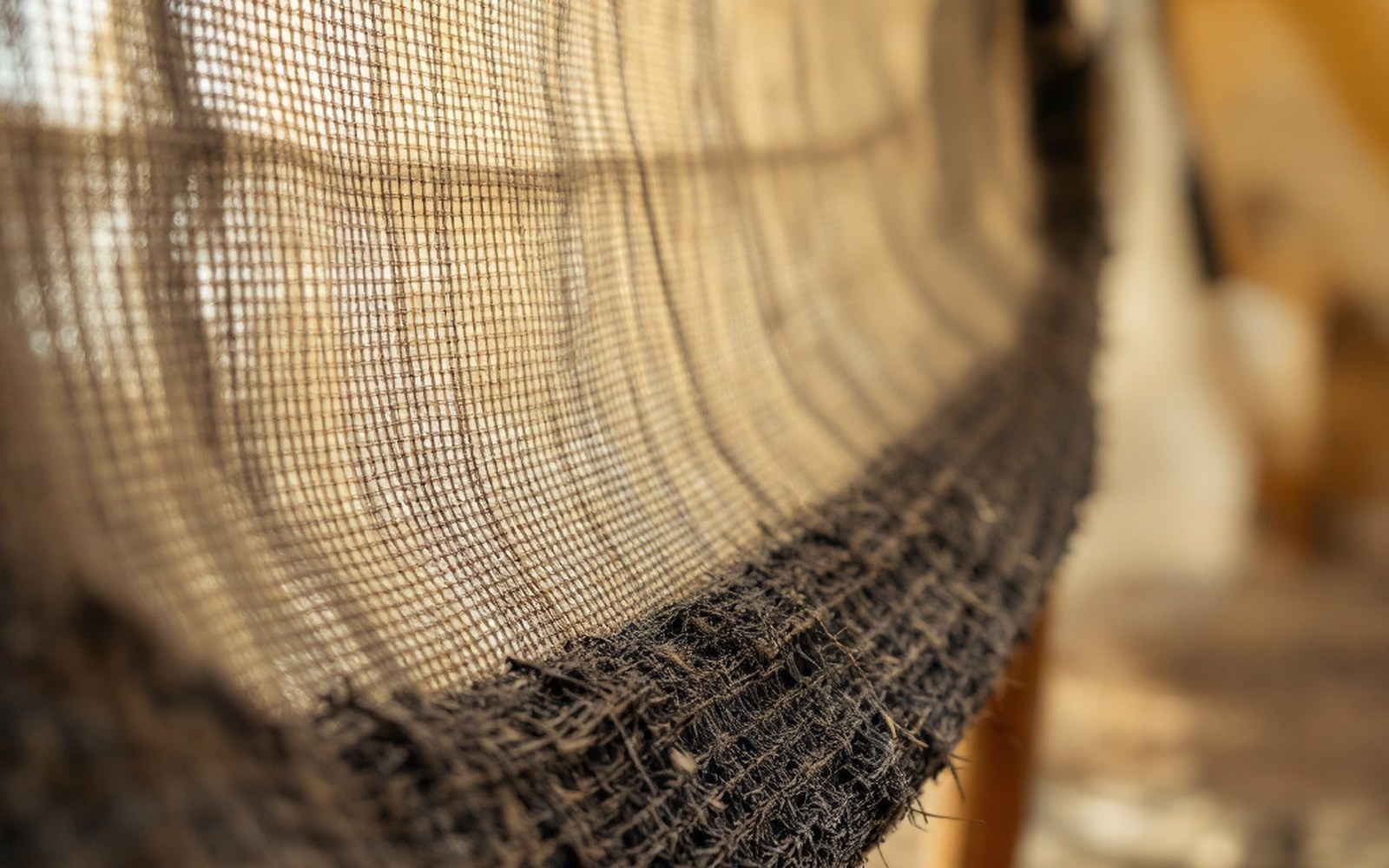 Close on coarse dark woven cloth stretched over a tent pole with daylight showing through the weave