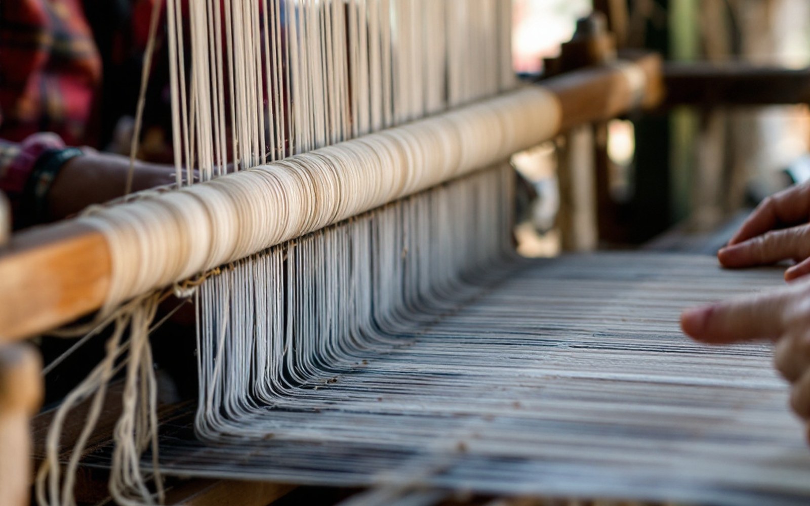 A narrow strip of woven woollen cloth on a simple loom with the warp under tension, adult hands at the shed